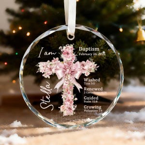 Könnte beinhalten: Runder, klarer Glasornament mit facettiertem Rand, mit einem Kreuzdesign, das mit rosa Blumen und einer rosa Schleife verziert ist. Der Ornament enthält den Namen "Stella" und den Text "Baptism February 26, 2026". Ein weißes Band zum Aufhängen.