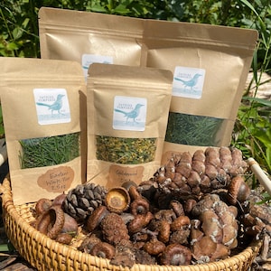 May include: Four brown paper bags filled with dried herbs and labeled with the text "Catbird Comforts". The bags are arranged on a woven wicker basket filled with acorns and pine cones.