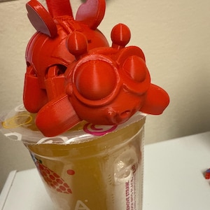 May include: A red 3D-printed character sits atop a clear plastic cup filled with a yellow beverage. The character has large, round eyes and a whimsical design. The cup has text that reads "PULL TAB & REMOVE STRAW."