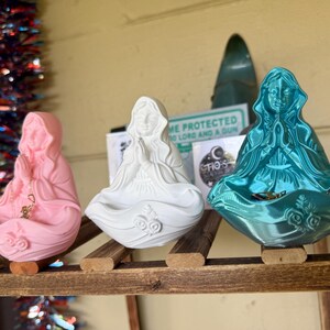 Può includere: Tre piccole statuette di una figura in preghiera, ognuna dotata di una base cava per riporre oggetti. Le statuette sono di colore rosa opaco, bianco opaco e con una finitura metallizzata color ottanio. Ogni figura ha le mani giunte e dettagli floreali.