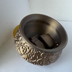 Op de afbeelding: Een bruine, decoratieve kom met een schedelpatroon aan de buitenkant. De binnenkant is donkerder bruin en de kom bevat twee rechthoekige bruine voorwerpen. De kom heeft een gele accentkleur aan de zijkant.