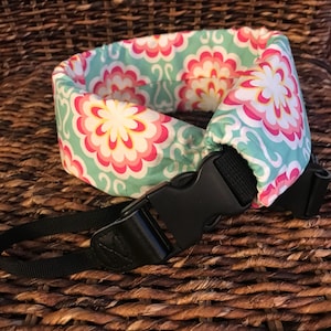 May include: A camera strap with a teal and pink floral pattern. The strap is padded and has a black adjustable buckle.