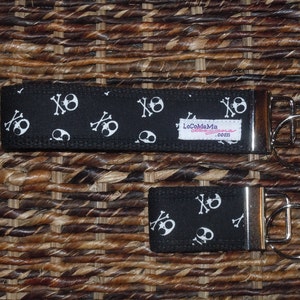 May include: Two black and white skull and crossbones patterned keychains with silver key rings. The keychains are made of fabric and have a black background with white skulls and crossbones. The text "LocoMama Designs.com" is printed on the larger keychain.