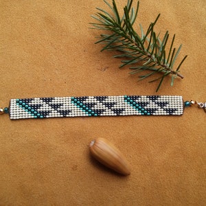 May include: A black, white, and turquoise beaded bracelet with a geometric pattern. The bracelet has gold-toned metal clasps.