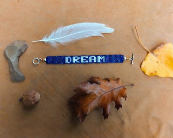 May include: A blue beaded bracelet with the word "DREAM" in white. The image also features a white feather, a yellow leaf, a brown leaf, a small acorn, and a stone object, all arranged on a brown surface.