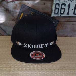 Könnte beinhalten: Schwarze Baseballkappe mit einem weißen, gestickten Band, auf dem "SKODEN" steht.
