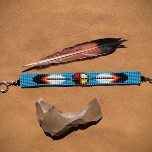 Può includere: Un braccialetto di perline turchesi con un designo nativo americano che presenta due piume e una ruota di medicina. Il braccialetto ha chiusure dorate ed è mostrato su una superficie marrone con una piuma e una pietra.