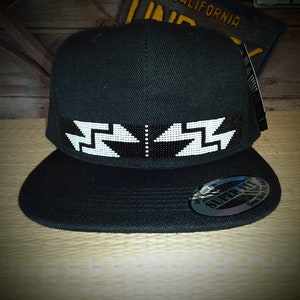 Peut inclure: Casquette de baseball noire avec un motif en perles blanches sur le devant. Le motif présente un motif géométrique avec des flèches pointant dans des directions opposées. La casquette a une visière noire et une sangle réglable noire.