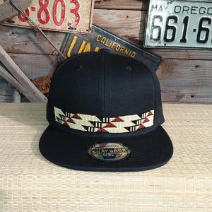 Könnte beinhalten: Schwarze Snapback-Kappe mit flachem Schirm. Die Kappe hat ein dekoratives Band mit einem geometrischen Muster in Rot, Weiß und Schwarz. Auf dem Schirm befindet sich ein Aufkleber.