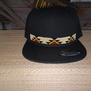 May include: Black baseball cap with a beaded band featuring a geometric pattern in white, yellow, and red. The band is labelled "Snapback".