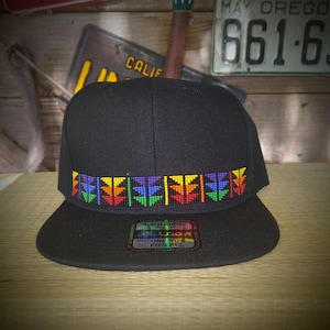 May include: A black baseball cap with a rainbow-colored beaded design around the crown. The cap has a curved brim and a black adjustable strap with a plastic buckle. The label on the strap reads "Legacy" and "One Size Fits All".