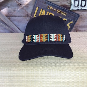 May include: A dark navy blue baseball cap with a decorative band of colorful beads. The beaded band features a geometric pattern with shades of turquoise, orange, and white. The cap is sitting on a light-colored surface.