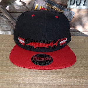 May include: Black and red snapback hat with a red bill. The hat features a red beaded design of a shark and white teeth. The hat has a snapback closure and a red button on top.