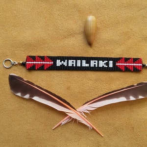May include: A black beaded bracelet with red and white accents. The bracelet has the word "WAILAKI" beaded in white letters. The bracelet is on a brown surface with two feathers and a small wooden object.