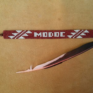 May include: A red beaded bracelet with white geometric patterns and the word "MODOC" in white beads. The bracelet has silver clasps on each end and is lying on a brown surface next to a brown and white feather.