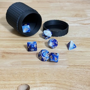 Puede incluir: Un conjunto de dados poliédricos azules y blancos sobre una superficie de madera. Los dados, de varias formas (d4, d6, d8, d10, d12, d20), están acompañados de un cubilete y una tapa negros. Los números de los dados son dorados.