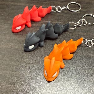 May include: Three dragon-shaped keychains in red, black, and orange. Each keychain features a detailed dragon design with white accents and a silver keyring. The keychains are made of a solid material and are suitable for attaching to keys or bags.