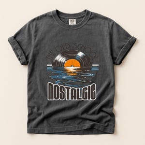 以下が含まれることがあります： 袖をまくった濃いグレーのTシャツ。水面に映るレコードのグラフィックで、「THE OLD SCHOOL」の文字が上に、「NOSTALGIC」の文字が下に配置されています。レトロな雰囲気のTシャツです。