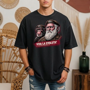 Peut inclure: Un t-shirt noir à manches courtes avec une impression graphique représentant un chimpanzé et un homme barbu portant des bérets assortis. Sous les personnages, le texte VIVA LA EVOLUTIC est imprimé en lettres blanches sur un fond rectangulaire rouge.