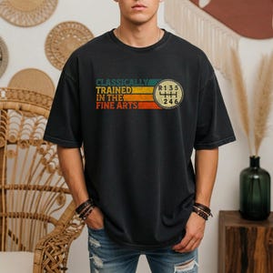 Może przedstawiać: Czarny t-shirt z krótkim rękawem z grafiką w poziome paski w kolorach morskim, żółtym, pomarańczowym i czerwonym. Napis "CLASSICALLY TRAINED IN THE FINE ARTS" znajduje się obok okrągłego schematu ręcznej skrzyni biegów.