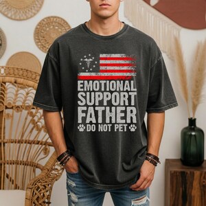 Può includere: T-shirt grigio scuro con un design a bandiera americana invecchiata e la scritta bianca "EMOTIONAL SUPPORT FATHER DO NOT PET". La maglietta ha un girocollo e maniche corte.