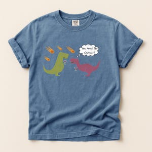 May include: Blue Comfort Colors t-shirt featuring a humorous graphic of two dinosaurs with wine glasses, observing falling meteors. One dinosaur says, "How About This Weather!?" in a speech bubble. The shirt has rolled-up sleeves.