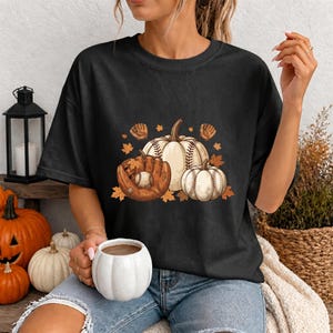 Peut inclure: Un t-shirt noir à manches courtes avec une impression graphique représentant deux citrouilles blanches aux coutures style baseball, un gant de baseball en cuir marron, une balle de baseball et des feuilles d'automne orange éparpillées.