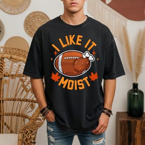 May include: A black short-sleeve t-shirt featuring a graphic of a brown football shaped like a roasted turkey. The text "I LIKE IT MOIST" is printed in orange above and below the graphic, accompanied by orange maple leaf and star accents.