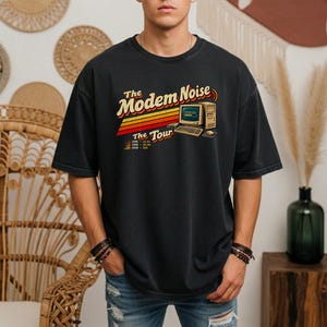 May include: A charcoal gray t-shirt featuring a retro graphic of a vintage computer with the text The Modem Noise The Tour. The design includes orange and yellow horizontal stripes and a list of years and connection speeds below the computer.
