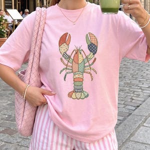 Może przedstawiać: Jasnoróżowy t-shirt z krótkim rękawem z centralną grafiką homara złożoną z różnych kolorowych wzorów patchworkowych, w tym motywów kwiatowych, w kropki i w kratę. Koszulka jest zestawiona z szortami w różowo-białe pionowe paski.