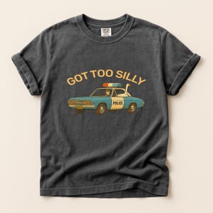 May include: Dark gray Comfort Colors t-shirt featuring a cartoon police car with a goose in the driver's seat. The text "GOT TOO SILLY" is above the car. The car is blue and orange with the word "POLICE" on the side.