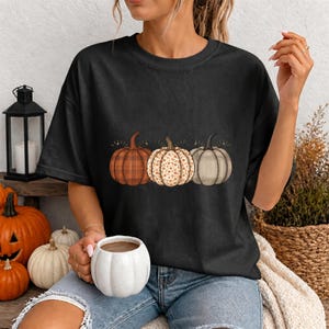 May include: A black short-sleeved t-shirt featuring a graphic print of three pumpkins arranged in a row. The left pumpkin has an orange plaid pattern, the centre pumpkin has a white and orange floral print, and the right pumpkin has a beige plaid pattern.