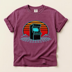 Puede incluir: Una camiseta granate con un diseño retro de videojuego arcade. El diseño incluye una máquina recreativa negra con una pantalla azul, sobre un fondo de puesta de sol con tonos naranja y rojo, y una cuadrícula azul. La camiseta tiene mangas enrolladas.