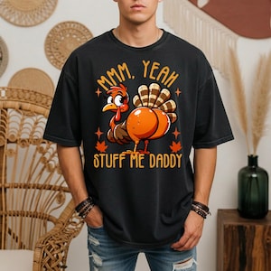 May include: A black short-sleeve t-shirt featuring a graphic of a cartoon turkey with orange and brown feathers. The text "MMM, YEAH" appears above the turkey, and "STUFF ME DADDY" is printed below it in orange, accompanied by small orange maple leaves.