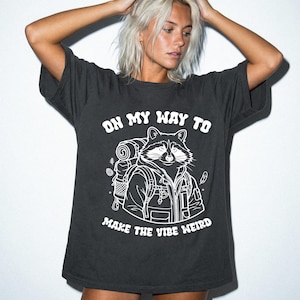 May include: Dark gray t-shirt with a white graphic of a raccoon wearing a backpack. The text "ON MY WAY TO MAKE THE VIBE WEIRD" is above and below the raccoon illustration. The shirt has short sleeves and a relaxed fit.