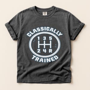 May include: Dark gray t-shirt with rolled-up sleeves. The shirt features a white and light blue graphic of a gear shift with the words "CLASSICALLY TRAINED" around it. The shirt is made of a soft, comfortable material.
