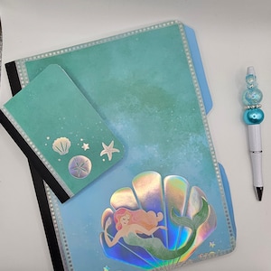 May include: A teal blue notebook with a smaller teal blue notebook on top. The larger notebook has a holographic mermaid design and the smaller notebook has seashells and starfish. A white pen with blue and iridescent beads is on the right side of the image.