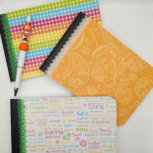 May include: Three small notebooks with colorful patterned covers. The covers feature a rainbow polka dot pattern, a paisley pattern, and a pattern with the words "You and Me", "Spirit", "Cherish", "Born to be Wild", "Life", "Full of", "Memories", "Golden Days", "Beautiful", "Forever", "Love", "Sweet", "Happy", "Imagine", "Together", "Cherished Moments", and "Beautiful".