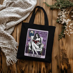 May include: Black tote bag featuring a tarot card design. The card depicts a woman reading a book, with the text "THE READER" at the bottom. The design includes bats, a pentagram, and a moon.