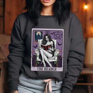 Puede incluir: Sudadera gris oscuro con un diseño de carta del tarot. La carta representa a una mujer leyendo un libro, con el texto "THE READER" en la parte inferior. La carta tiene un borde púrpura con murciélagos y un pentagrama. La mujer está sentada en una silla blanca.