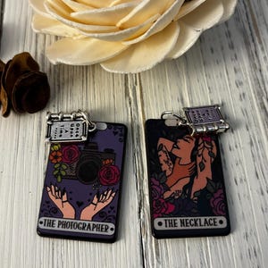 May include: Two purple and black tarot cards with gold accents. The cards are titled "The Photographer" and "The Necklace". Each card has a small charm attached to the top that reads "Once Upon A Time".