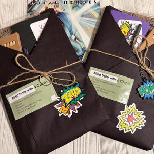 May include: Two black paper envelopes tied with twine, labeled "Blind Date with a Comic," each with a key chain and comic book-themed stickers. The contents include a comic, popcorn, notebook, coloring pencils, and a key chain. A comic book is visible in the background.