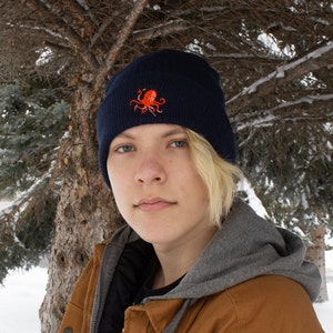 May include: A person wearing a navy blue beanie with an embroidered red octopus design. The beanie is pulled down over their forehead and they are wearing a brown jacket with a gray hoodie underneath.