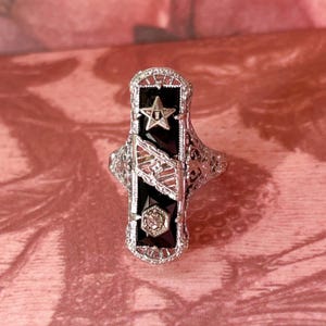 May include: A silver-toned ring with a rectangular black onyx center, featuring a star and geometric accents. The ring has intricate filigree detailing and a small, diamond-like stone. The design is vintage-inspired.