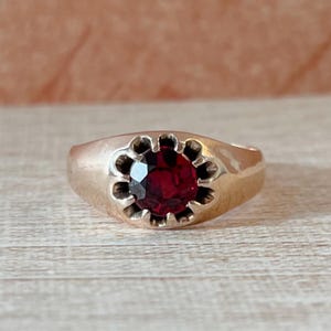 May include: A gold-toned ring featuring a large, round, deep red gemstone. The gemstone is set in a floral-shaped bezel. The ring has a smooth, polished band and a classic, elegant design.