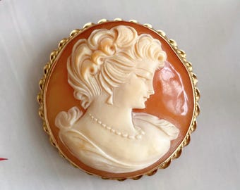 May include: A cameo brooch featuring a carved portrait of a woman with a detailed hairstyle. The cameo is set in a gold-toned metal frame with a scalloped edge.