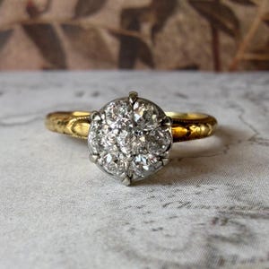 May include: A gold ring with a cluster of small, clear gemstones set in a circular pattern. The band has an ornate, engraved design. The ring is set against a neutral background, highlighting the sparkle of the gemstones.