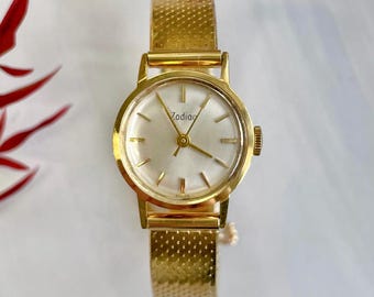 May include: A gold-toned Zodiac wristwatch with a white face and gold hands. The watch has a gold mesh band.