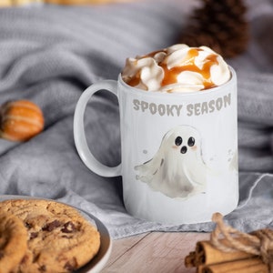 May include: White ceramic mug with a ghost graphic and the text "Spooky Season". The mug is filled with a hot beverage topped with whipped cream and caramel sauce.