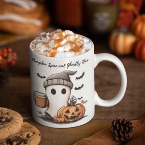 May include: White ceramic mug with a ghost holding a cup of coffee and a pumpkin. The mug has the text "Pumpkin Spice and Ghostly Nice" printed on it.